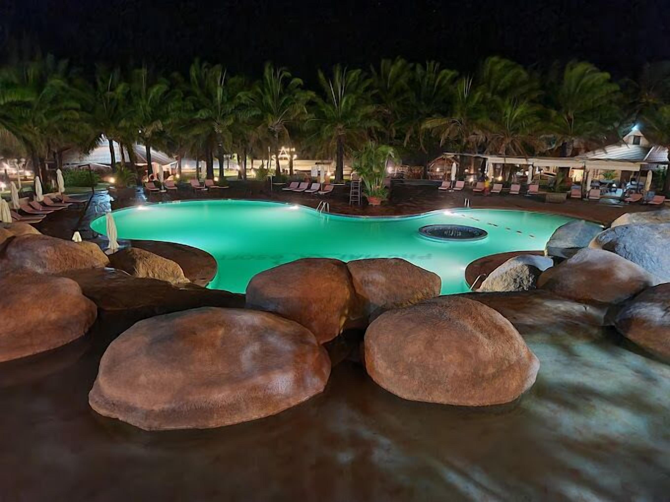A nighttime pool area with large rocks, lounge chairs, and palm trees.