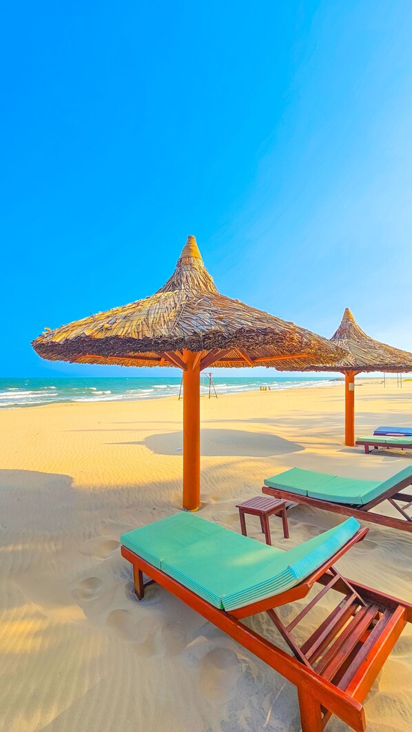 A beach with thatched umbrellas and wooden lounge chairs. -  Phu Hai Beach Resort & Spa Phan Thiet (Phan Thiet)