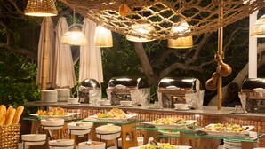 Montaje de bufé al aire libre con diferentes estaciones de comida, mobiliario de mimbre e iluminación colgante.