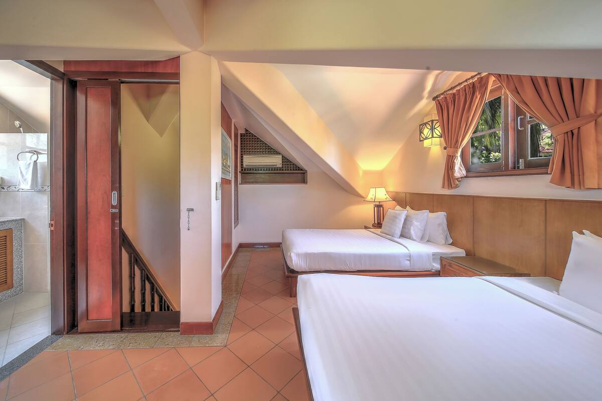 A hotel room with two beds, a window with curtains, a lamp, and a staircase leading to an upper level.