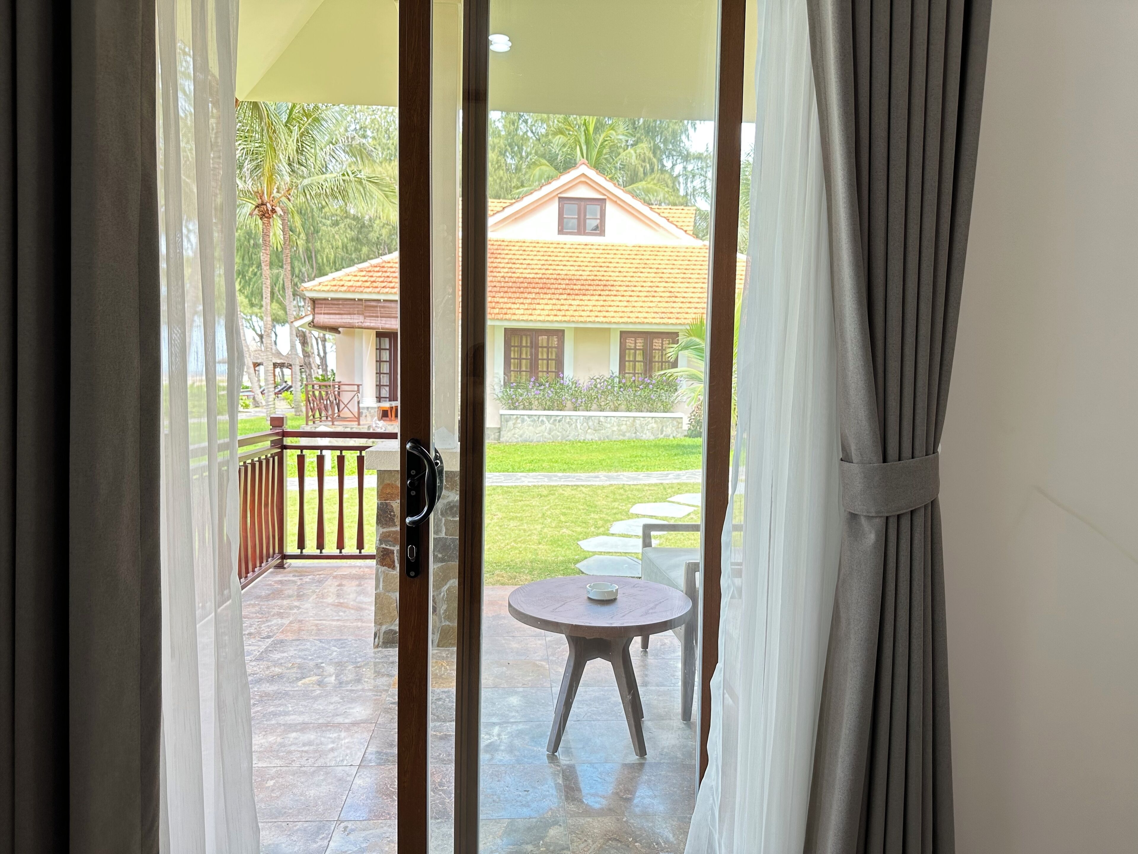 On aperçoit, à travers une porte-fenêtre coulissante, un espace extérieur aménagé en terrasse, avec une table et des chaises, un petit jardin et un bâtiment doté d’un toit en tuiles.