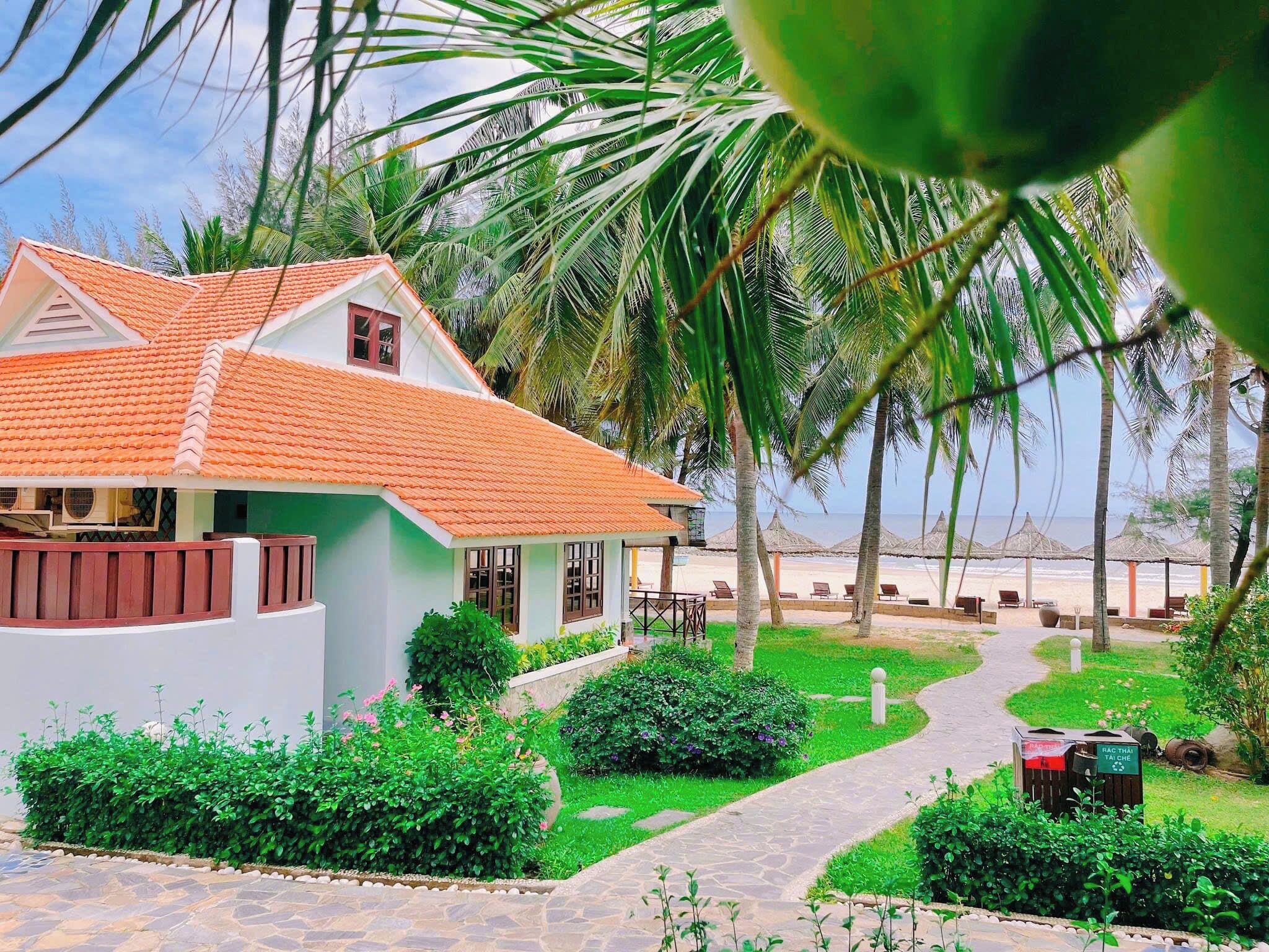 Un bungalow con tetto in tegole, circondato da vegetazione rigogliosa e un vialetto che conduce a una spiaggia con ombrelloni.