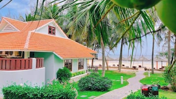 Un bungalow con tetto in tegole, circondato da vegetazione rigogliosa e un vialetto che conduce a una spiaggia con ombrelloni.