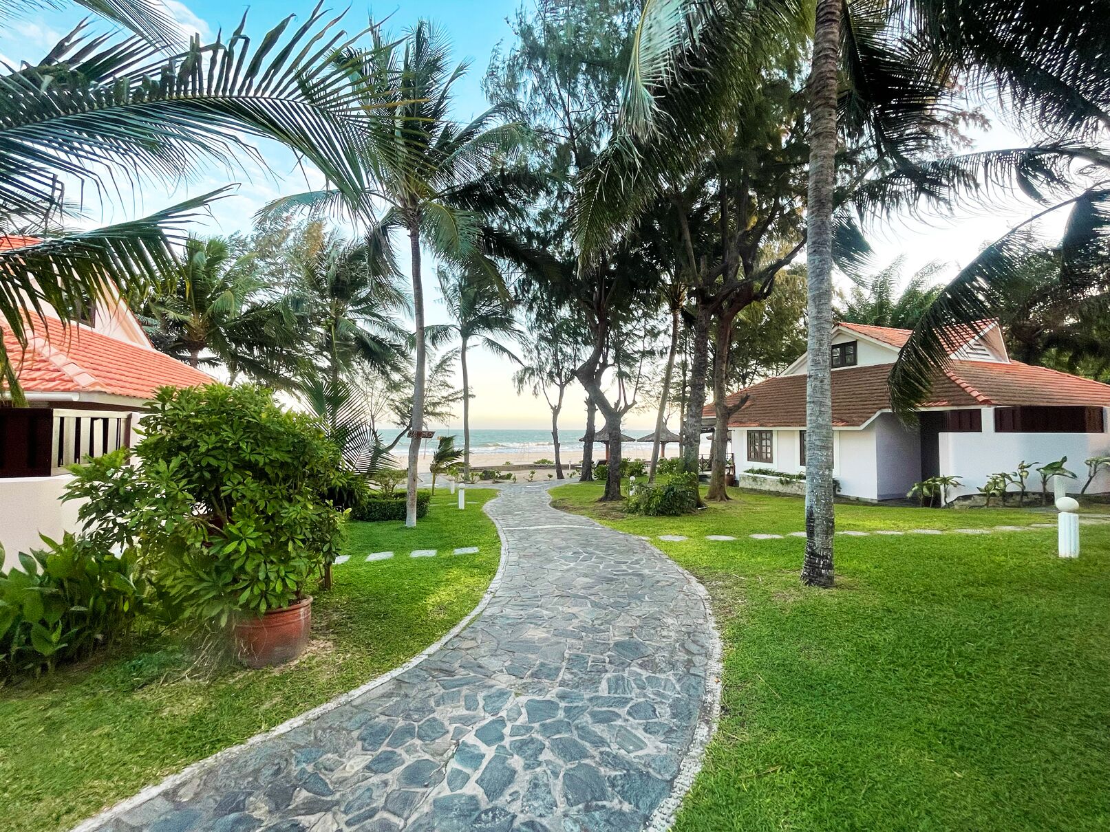 Ein Weg führt zu einem Strand mit Palmen und einem Gebäude mit rotem Dach.