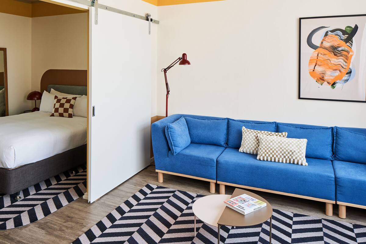 A modern hotel room with a bed, a blue sofa, a small round table, and a painting on the wall.