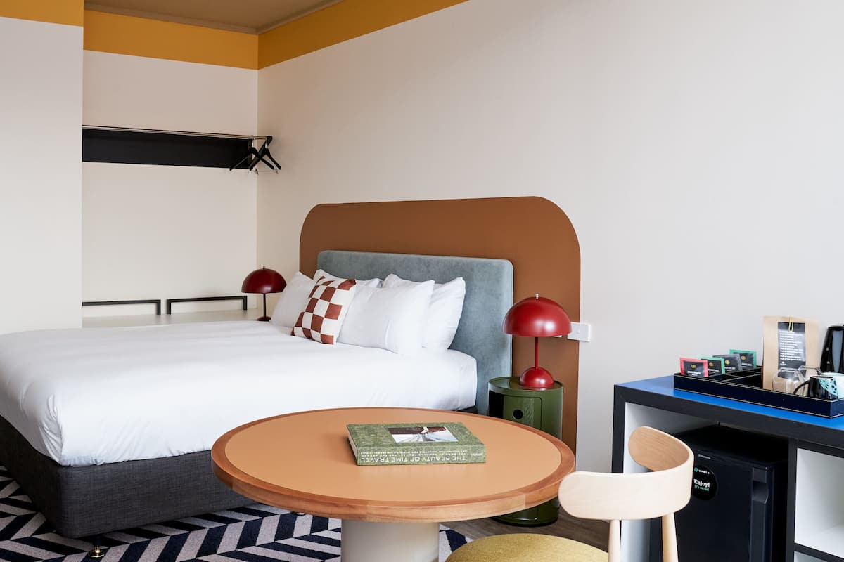 A hotel room with a bed, a round wooden table, a desk with a chair, and a small bookshelf.