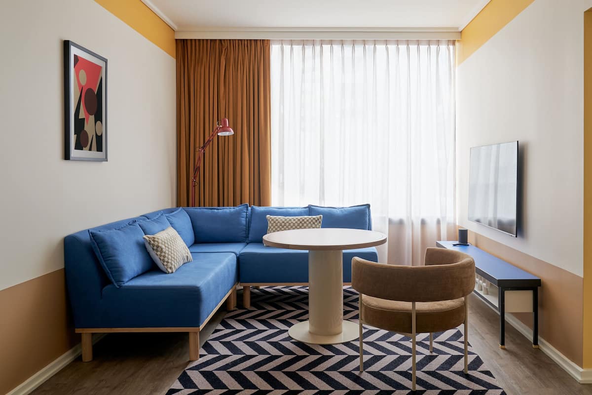 A modern hotel room with a blue sectional sofa, a round table, a brown armchair, a TV, and a patterned rug.