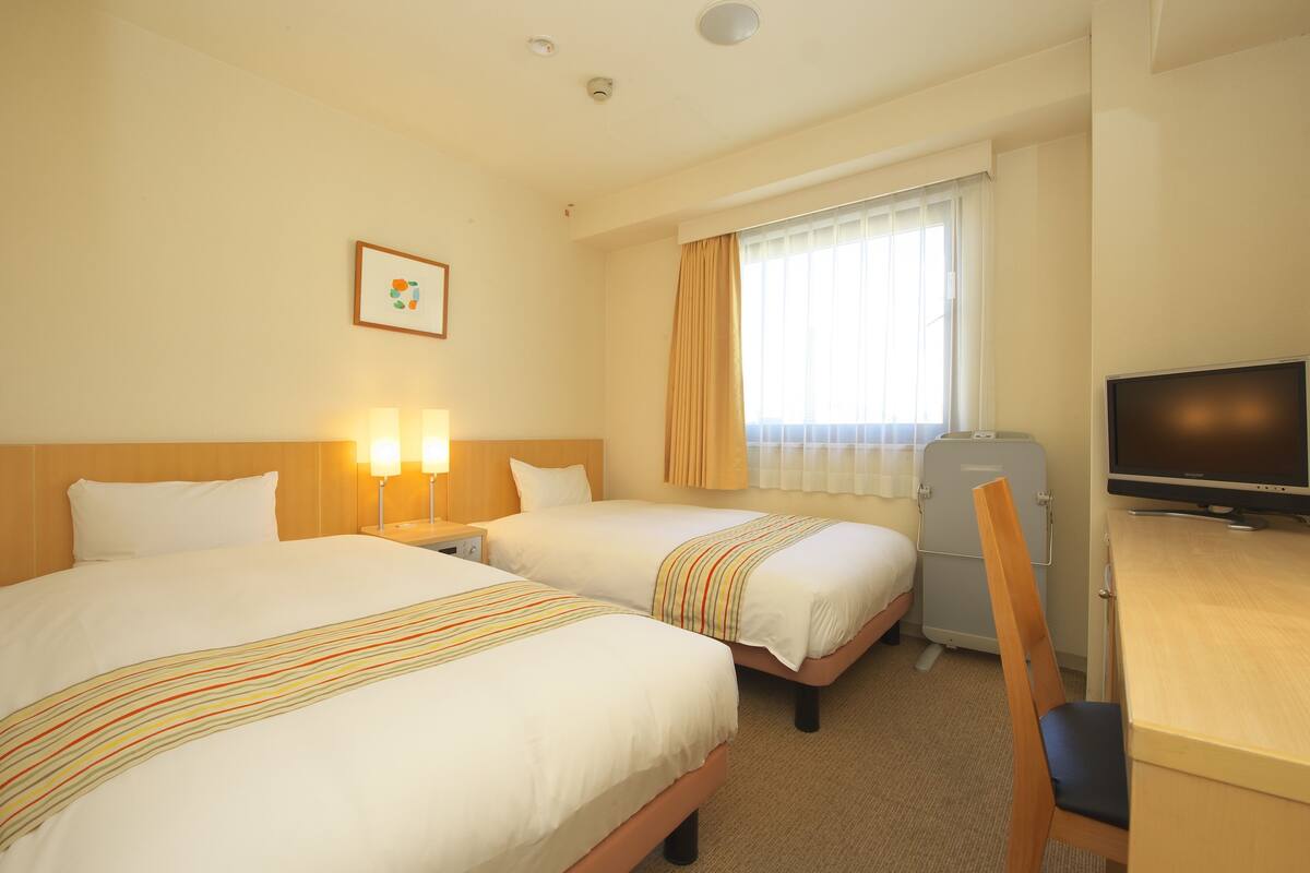 A hotel room with two beds, a desk, a television, and a window with curtains.