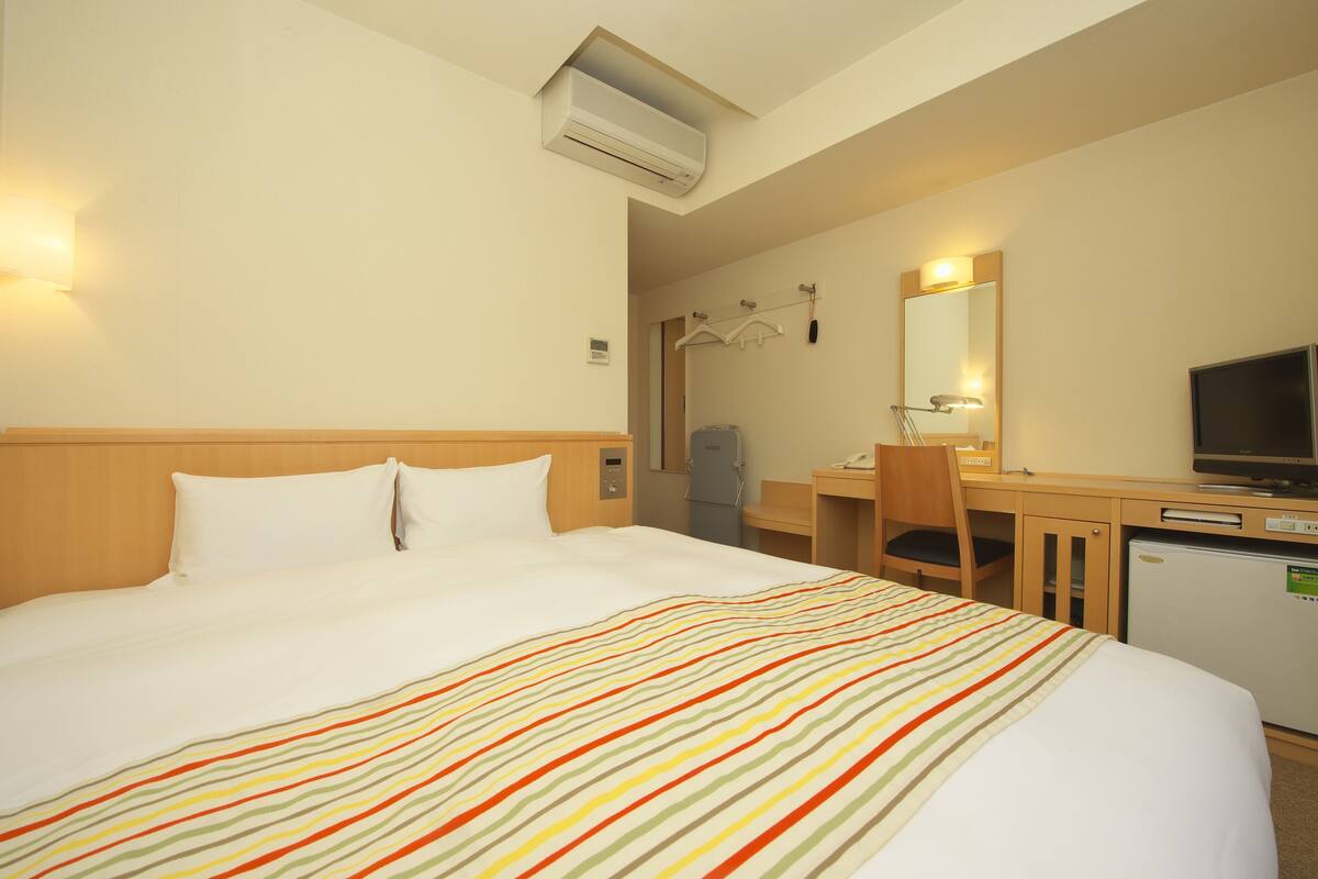 A hotel room with a bed, a desk, a chair, a television, and an air conditioning unit.