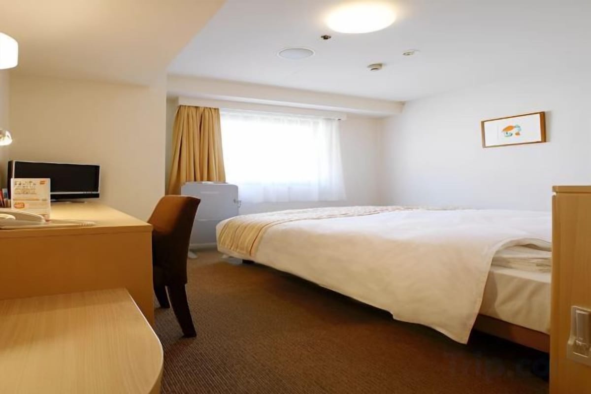 Una habitación de hotel con cama, escritorio con silla, televisor y ventana con cortinas.