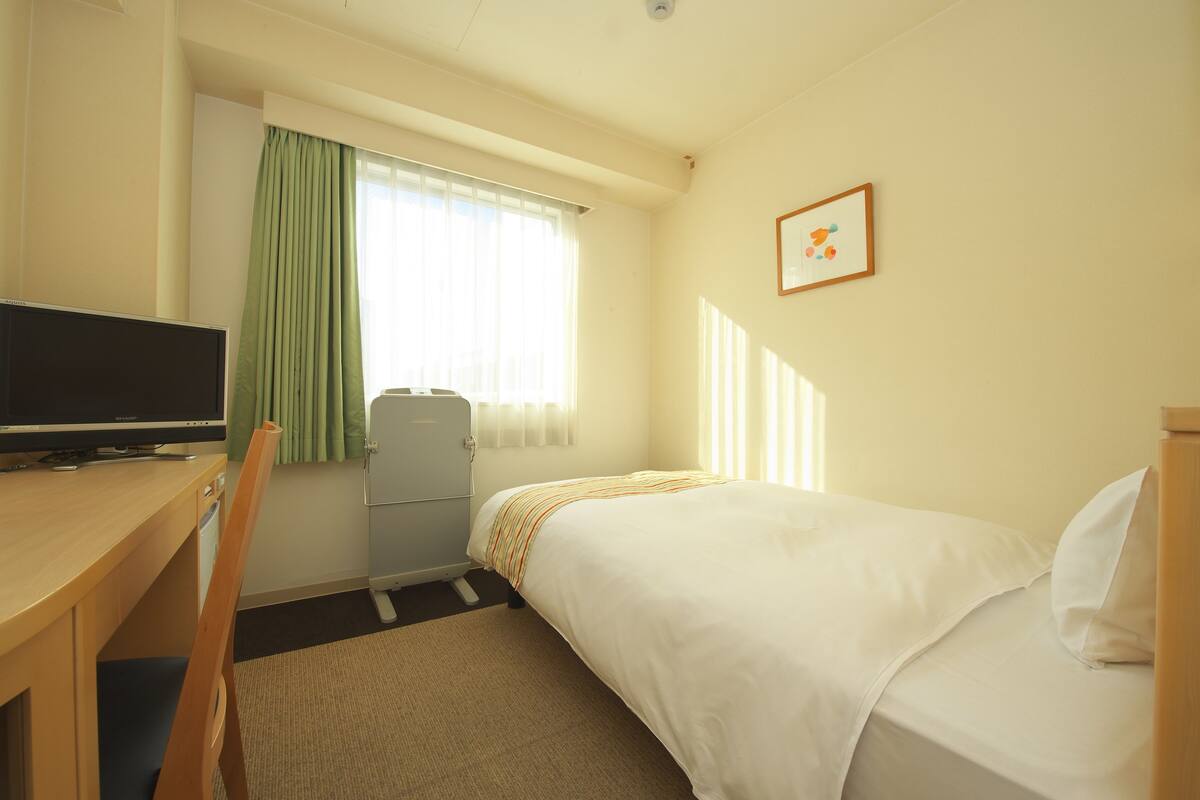 A hotel room with a bed, a desk with a television, a chair, and a window with curtains.
