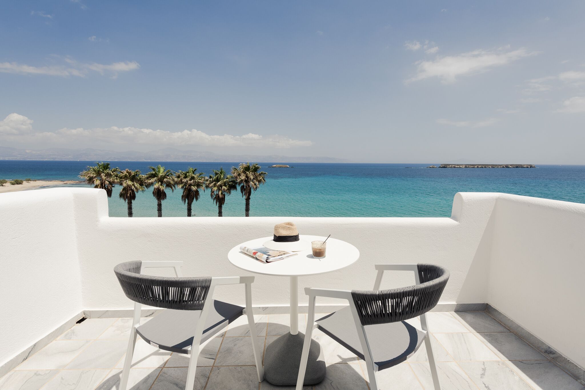 Un balcón con una mesa blanca y dos sillas, con vistas a una playa y al mar.