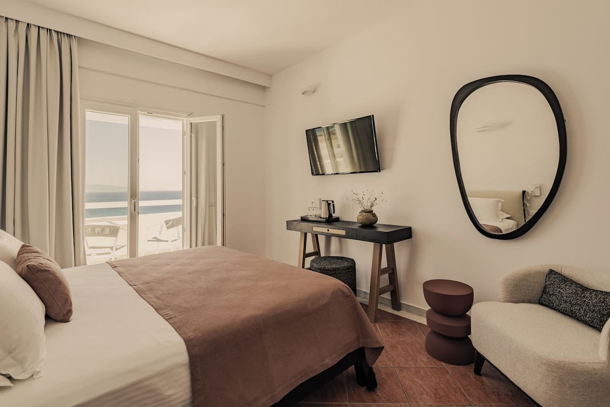 A hotel room with a bed, a desk with a flat-screen TV, a chair, and a view of the sea through large windows.