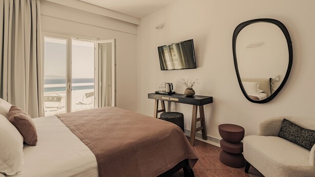 A hotel room with a bed, a desk with a flat-screen TV, a chair, and a view of the sea through large windows.