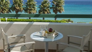 A balcony with wicker chairs, a table with a fruit bowl and two glasses, and a view of the ocean and palm trees.