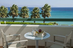 A balcony with wicker chairs, a table with a fruit bowl and two glasses, and a view of the ocean and palm trees.