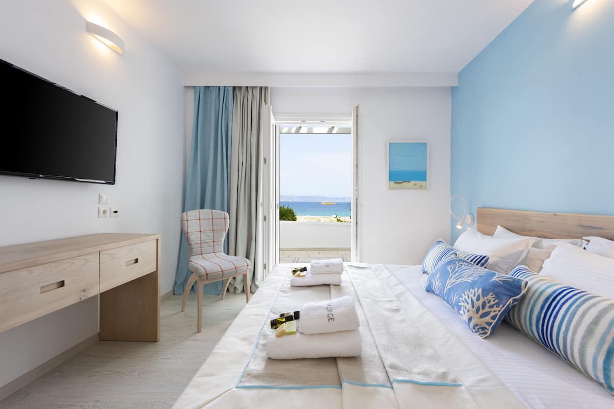 A hotel room with a bed, a desk, a chair, a TV, and a view of the beach.