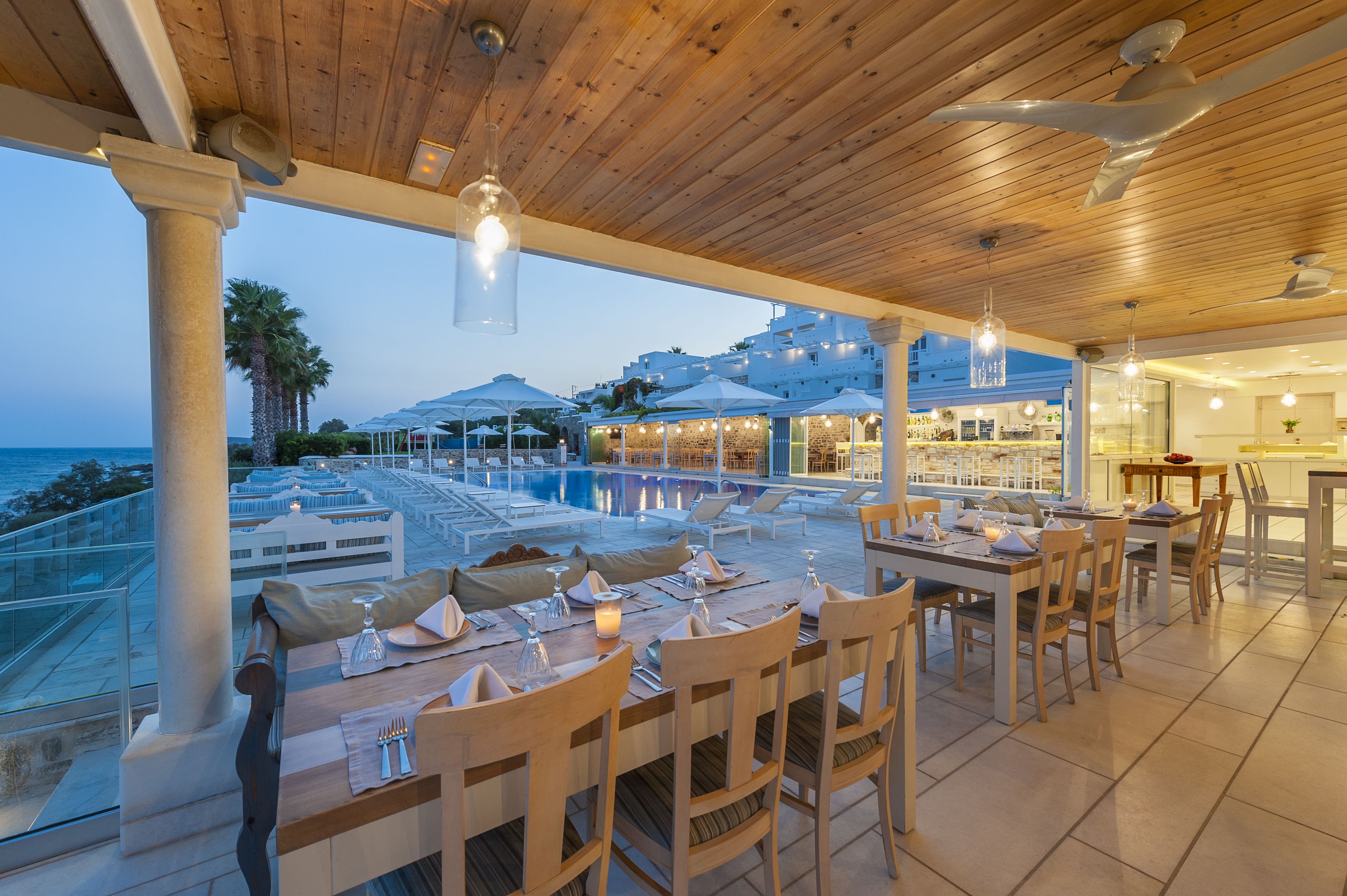 A rooftop dining area with tables set for two, overlooking a pool and the sea.