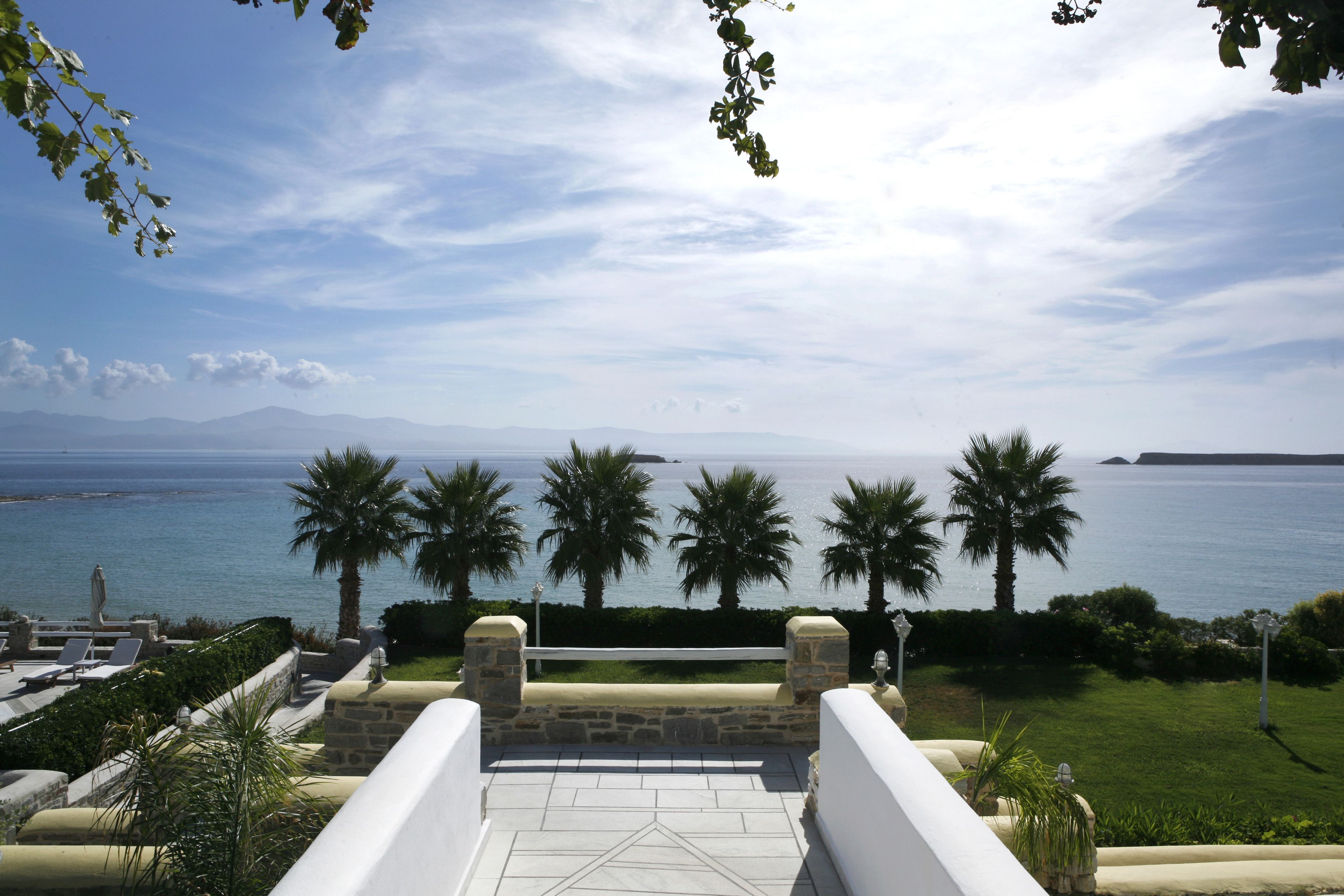 A terrace with a view of the sea, palm trees, and distant mountains.