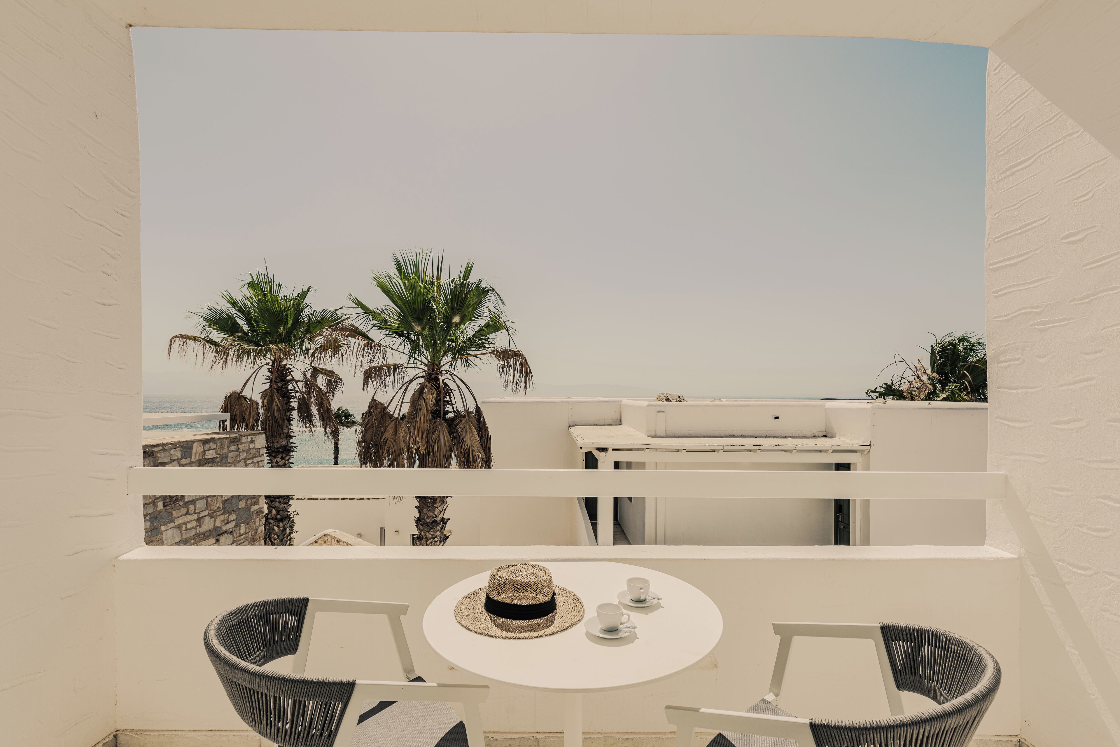 A balcony with a white table and two wicker chairs, overlooking a beach with palm trees and a clear sky.
