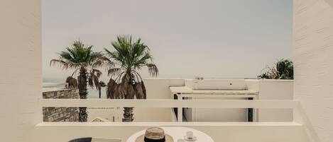 A balcony with a white table and two wicker chairs, overlooking a beach with palm trees and a clear sky.