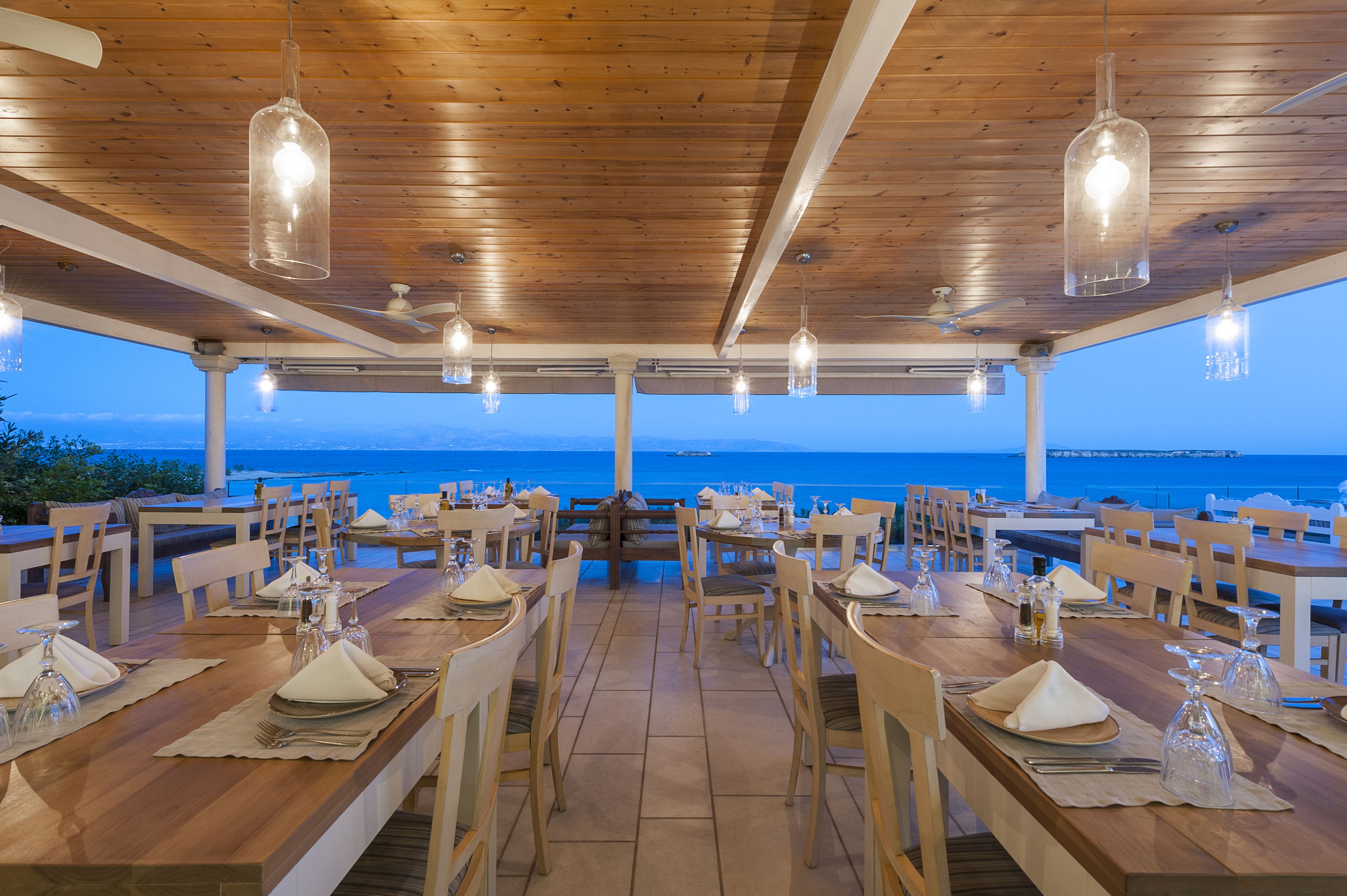 An outdoor dining area with wooden tables and chairs, set against a backdrop of the sea and mountains.