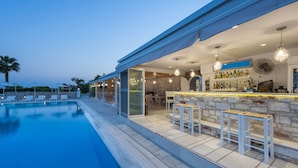 A poolside bar area with a stone wall, bar stools, and a variety of bottles. - Saint George Hotel (Paros)