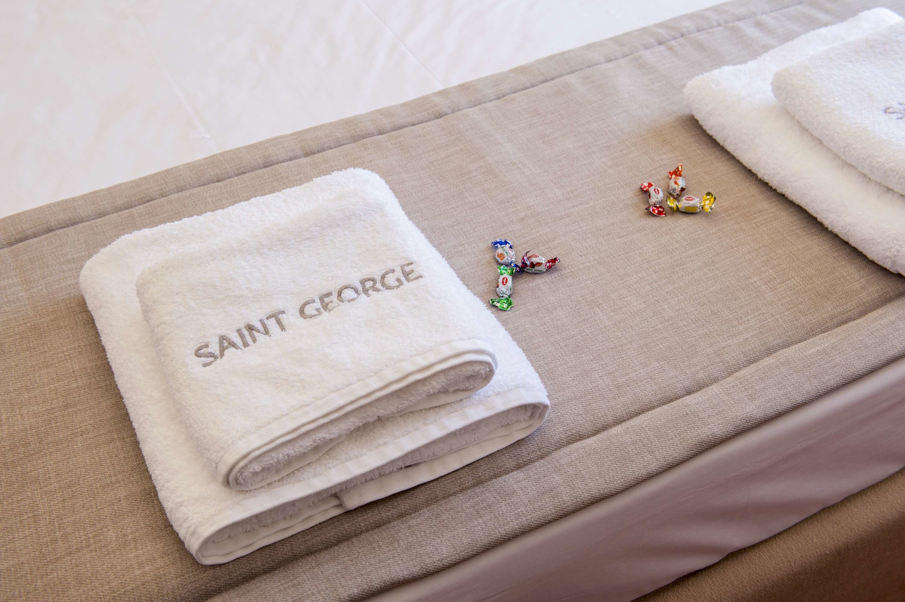 A neatly folded white towel with "SAINT GEORGE" embroidered on it, placed on a beige bedspread.