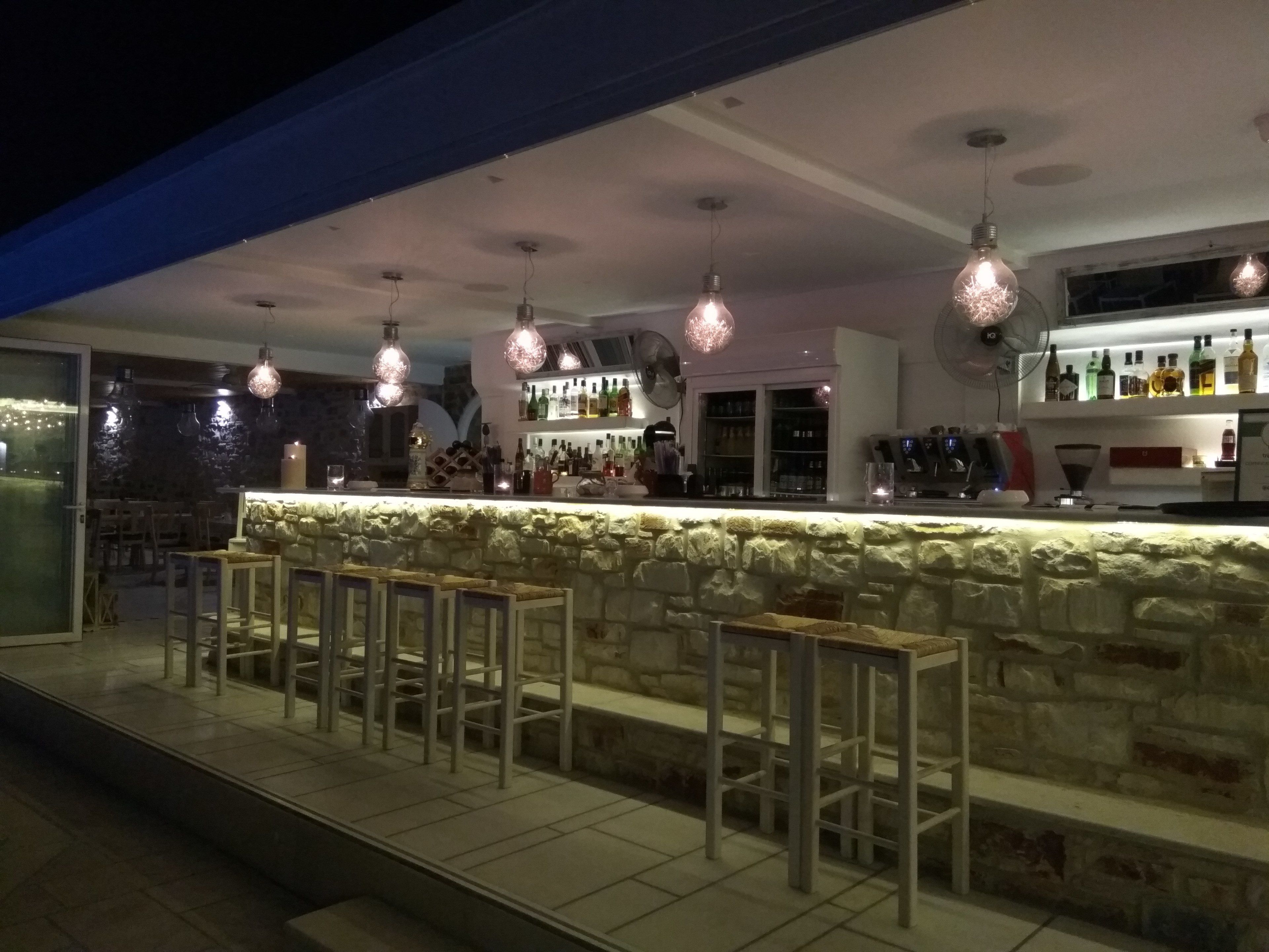 Zona de bar al aire libre con mostrador de piedra y taburetes, iluminación colgante y una variedad de botellas y vasos en estanterías.