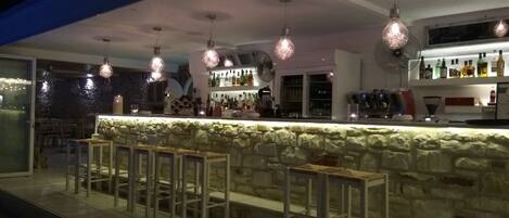 An outdoor bar area with stone counter and bar stools, overhead pendant lighting, and a variety of bottles and glasses on shelves.