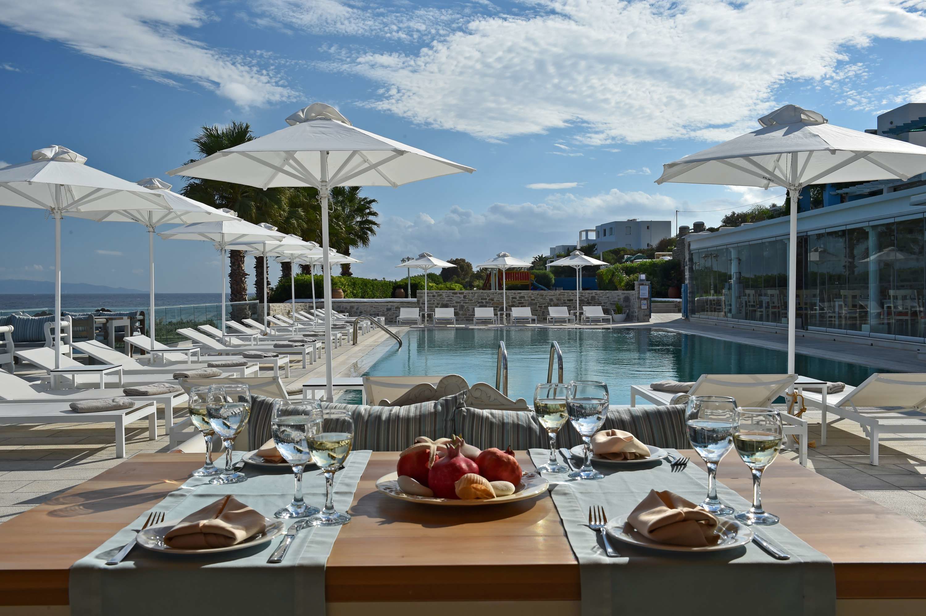 A poolside dining area with white umbrellas, lounge chairs, and a table set with plates of food and glasses of wine.