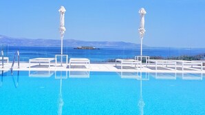 A clear blue pool with white lounge chairs and umbrellas, surrounded by a white fence, with a view of the sea and distant mountains.