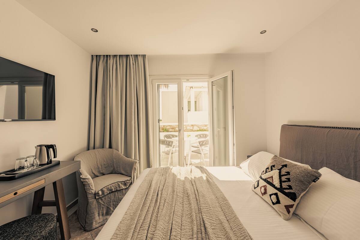 A hotel room with a bed, a desk with a kettle, a chair, and a view of the outside.