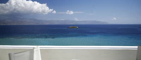 Un balcón con sillas y una mesa blancos, con vista a un mar tranquilo y una pequeña isla a lo lejos, con montañas al fondo.