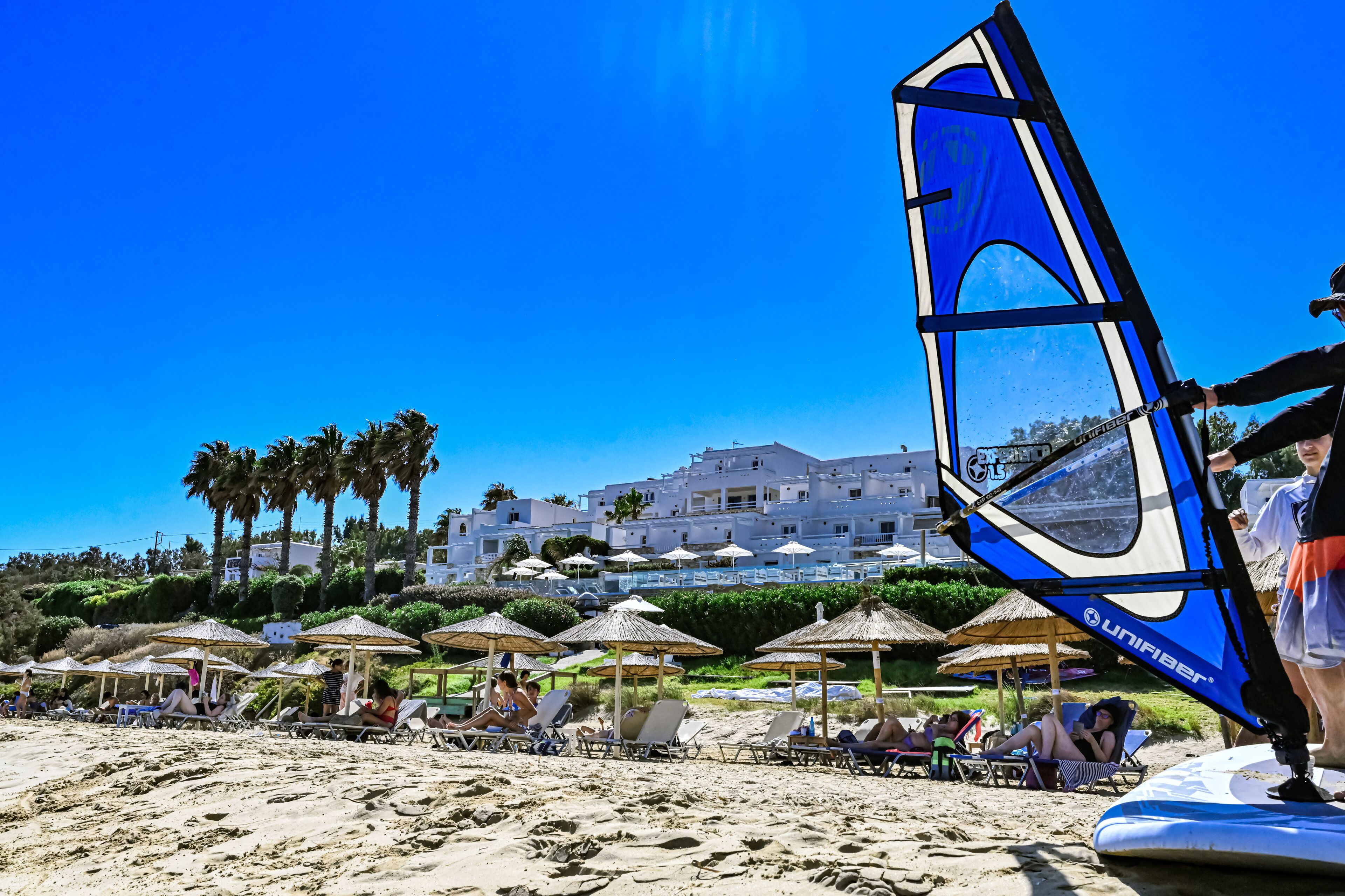 A beach with sun loungers and umbrellas, a windsurfing board, and a building in the background.