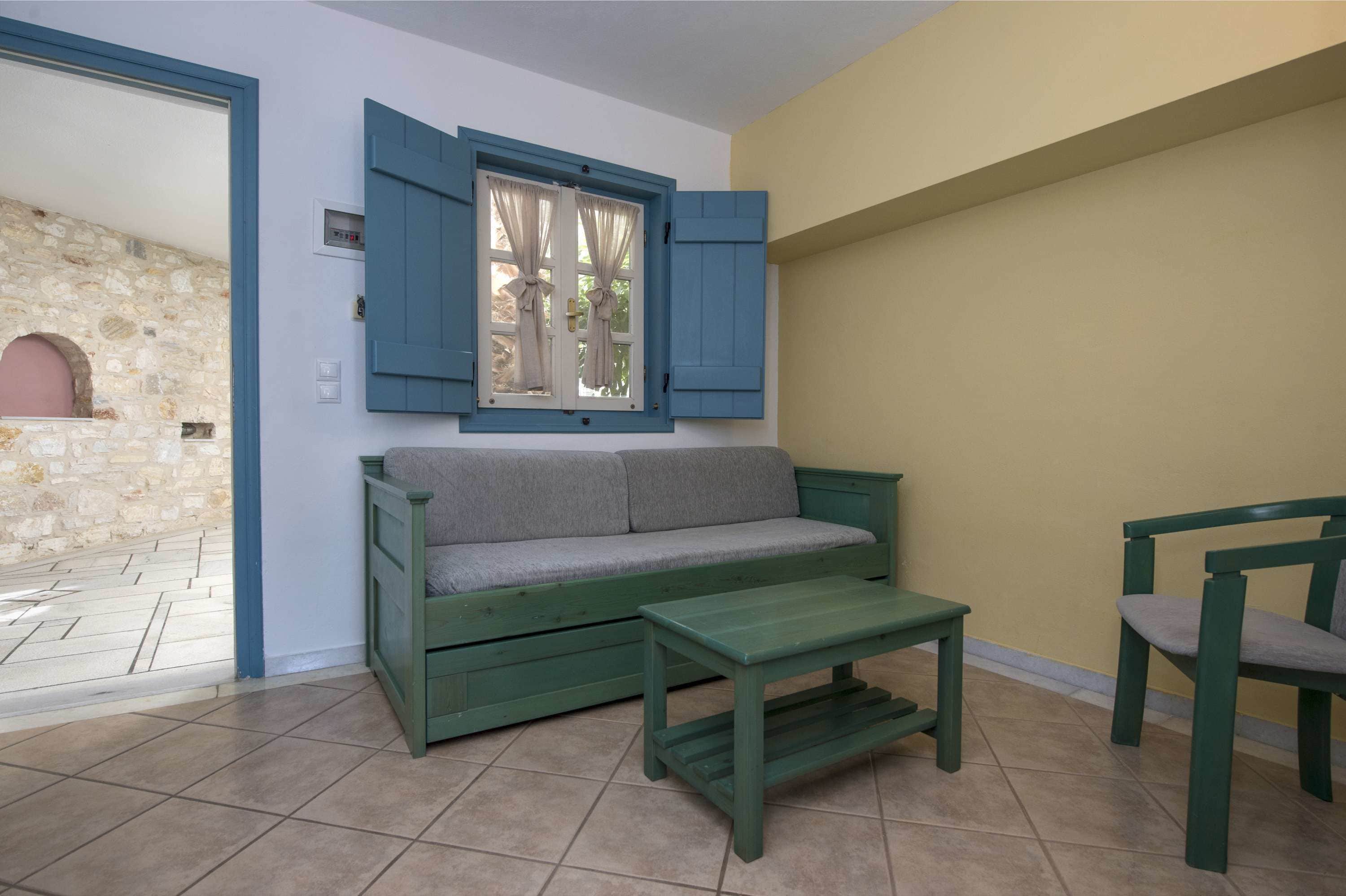 Quarto com sofá verde, mesa do mesmo tom e janela com persianas azuis.