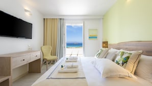 A hotel room with a bed, a desk, a chair, a TV, and a view of the sea. - Saint George Hotel (Paros)