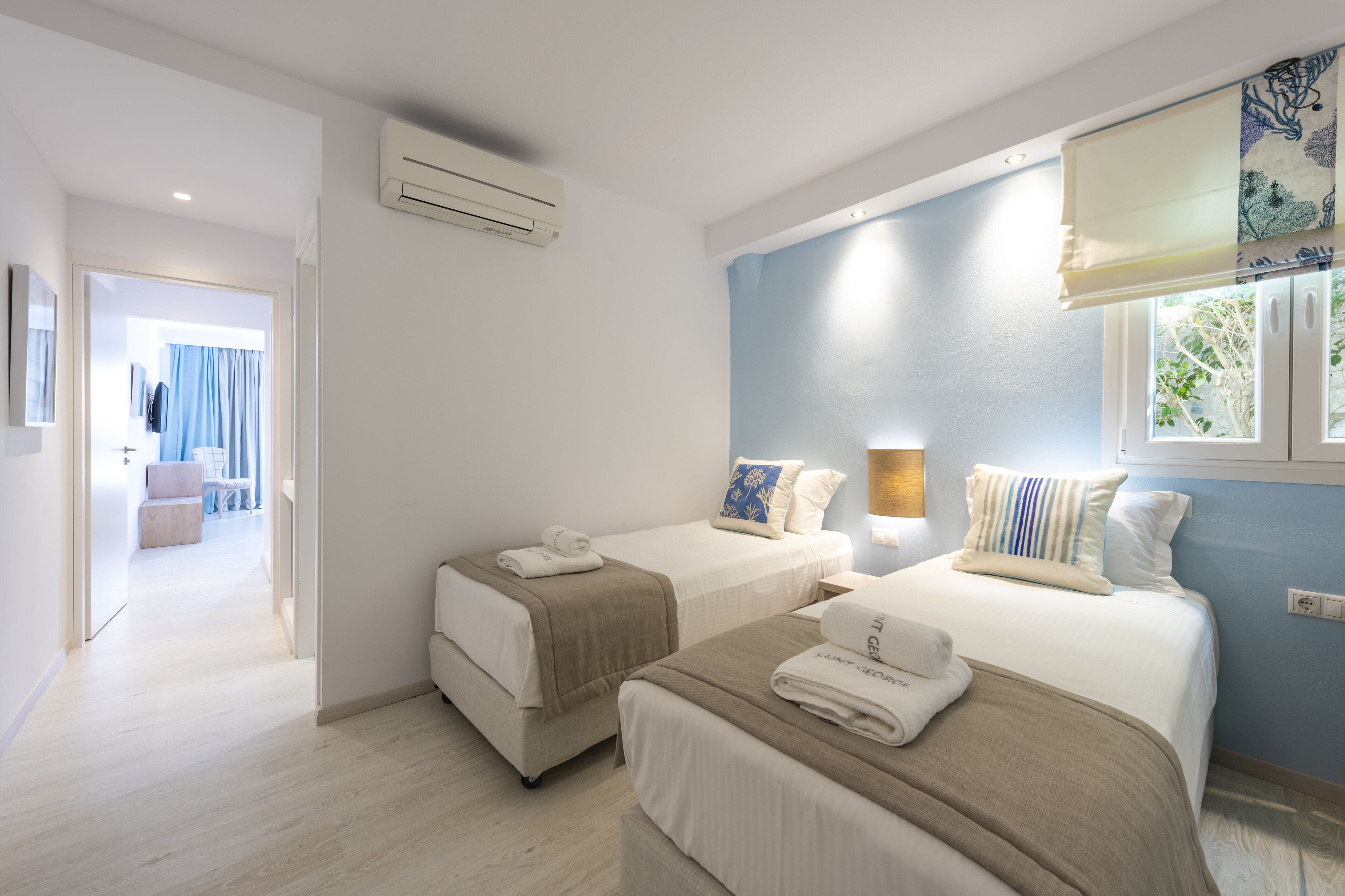 A hotel room with two beds, a window with curtains, an air conditioning unit, and a hallway leading to another room.