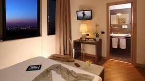 A hotel room with a bed, a desk with a lamp, a TV, and a view of the city at dusk. - Hotel & Spa Villa Mercede (Frascati)