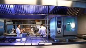A commercial kitchen with stainless steel appliances, two chefs working, and a walk-in refrigerator. - Hotel & Spa Villa Mercede (Frascati)