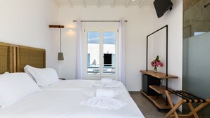 A hotel room with a large bed, a wooden headboard, a bedside table with a mirror, a small wooden shelf with a vase of flowers, and a view of the city through the window. - Magas Hotel (Mykonos)