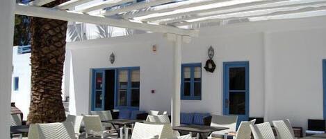 Un espacio al aire libre con sillas y mesas blancas, contraventanas azules y un edificio blanco con un pilar de piedra.