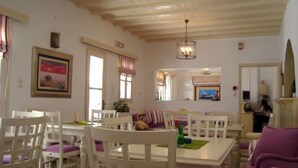 A dining area with white wooden chairs and a table, a ceiling with exposed beams, and a painting on the wall. - Magas Hotel (Mykonos)
