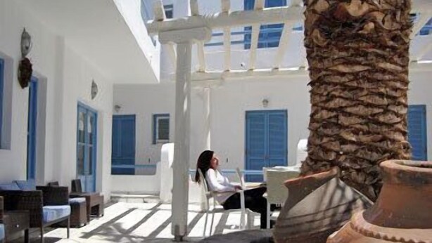 A white building with blue shutters, a palm tree, and outdoor seating. - Magas Hotel (Mykonos)