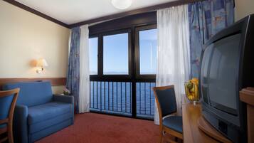 Habitación de hotel con sofá, una silla, un televisor y vista al mar a través de amplias ventanas.