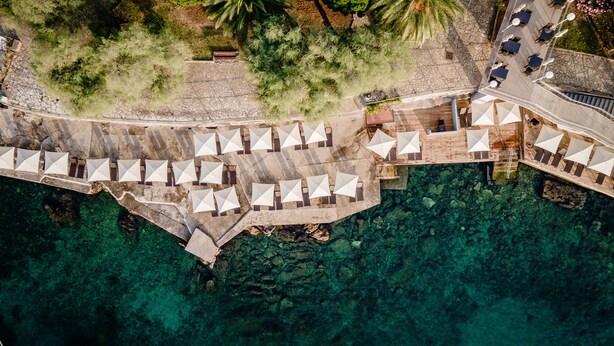 An aerial view of a coastal hotel with a series of outdoor umbrellas and seating areas along the waterfront, a stone pathway, and a wooden deck with tables and chairs. - Hotel Kristal - Liburnia (Opatija)