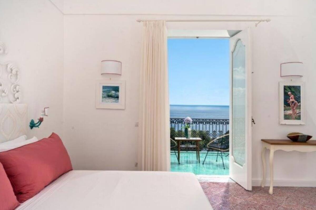 A bedroom with a bed, a small table, and a view of the sea through an open door.