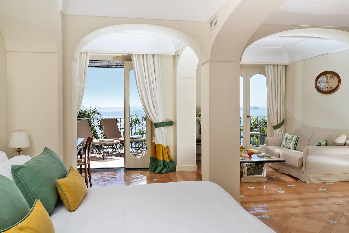 A hotel room with a large bed, a sofa, a dining table, and a view of the sea.