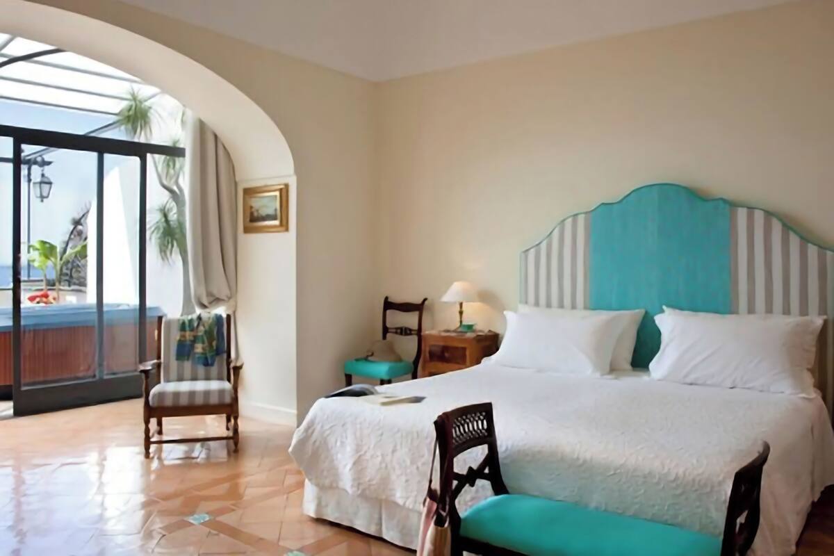 A hotel room with a bed, a chair, a nightstand, a lamp, and a view of the outdoors through a glass door.
