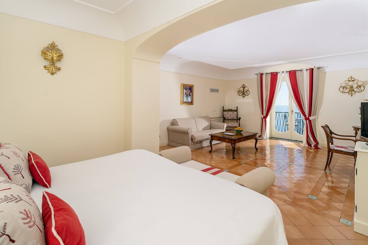 A hotel room with a large bed, a sofa, a dining table, and a view of the balcony.